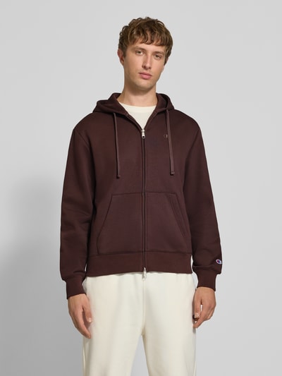 Männermodel trägt Sweatjacke mit Label-Stitching und Känguru-Tasche von Champion im Baumwoll-Mix in Dunkelbraun, Ganzkörperansicht von vorne.