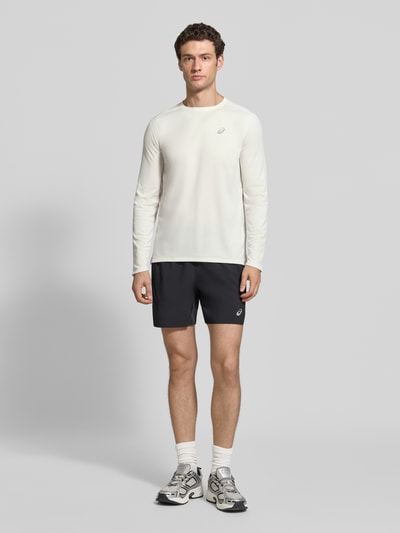 Männermodel trägt Regular Fit Langarmshirt mit Logo-Print von ASICS in Weiß, Rundhalsausschnitt, schwarze Shorts, neutrale Farben, Ganzkörperansicht von vorne. Rückenlänge bei Größe M: 72 cm.