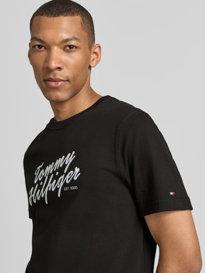 Männermodel trägt Regular Fit T-Shirt aus reiner Baumwolle von Tommy Hilfiger in Unifarbe Schwarz mit Label Print, Nahaufnahme Oberkörper.
