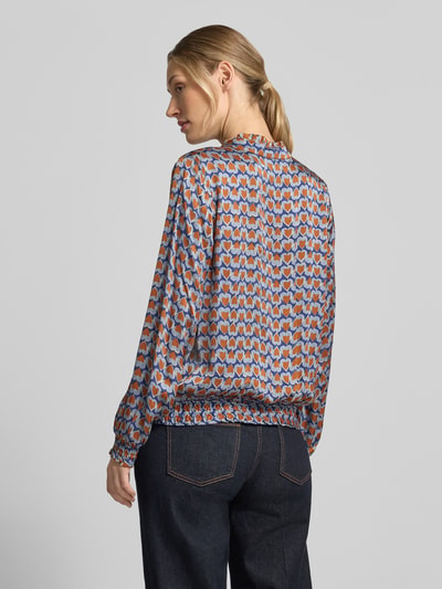 Vrouwenmodel draagt Regular Fit blouse uit zuiver viscose van MORE, abstract patroon met blauw, oranje en wit, rugaanzicht. Ruglengte maat 36: 57 cm, mouwlengte maat 36: 59 cm, opstaande kraag met ruches, v-hals, blouse met lange mouwen, elastische mouwboordjes.