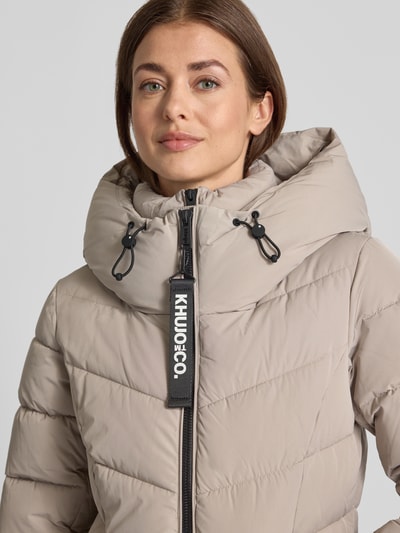 Vrouwenmodel draagt taupekleurig gewatteerd jack met capuchon en labeldetail, Model 'HERSCH2' van khujo, doorgestikte naden, recht model, praktische ritssluiting, zij-ritszakken, gebreide mouwboorden, gladde textuur, Nahaufnahme bovenlichaam.