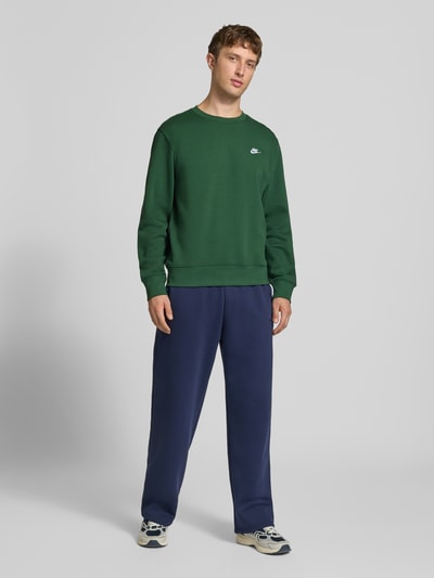 Männermodel trägt Relaxed Fit Sweatshirt aus Baumwoll-Mix von Nike in Dunkelgrün, kombiniert mit dunkelblauer Hose, Ganzkörperansicht von vorne. Herren Sweatshirt von Nike.