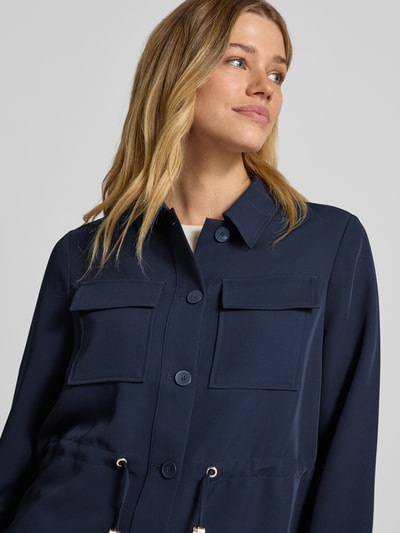 Kobiece modelka ma na sobie Regular Fit Longblazer mit Brustpattentaschen od s.Oliver BLACK LABEL, kolor granatowy, gładka faktura, dwie kieszenie z patkami na piersi, kołnierz klasyczny, zapięcie na guziki, tunel w talii, długie rękawy z mankietami, widoczny prosty krój regular fit, zdjęcie: zbliżenie na górną część ciała.