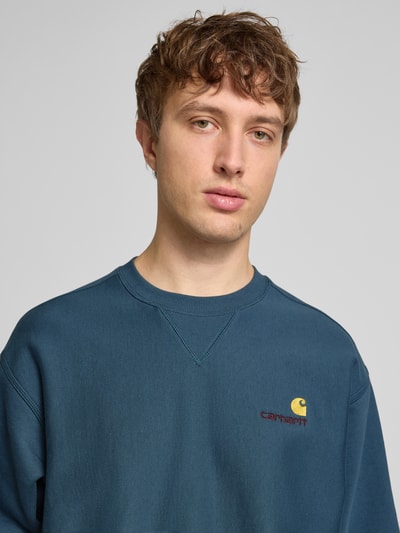 Męski model nosi bluza Sweatshirt mit Label-Stitching od Carhartt Work In Progress, mieszanka bawełny, luźny krój, prążkowane wykończenia, okrągły dekolt, wyhaftowane logo, kolor niebieski, długość tyłu 69 cm, długość rękawa 65 cm, zbliżenie na górną część ciała.