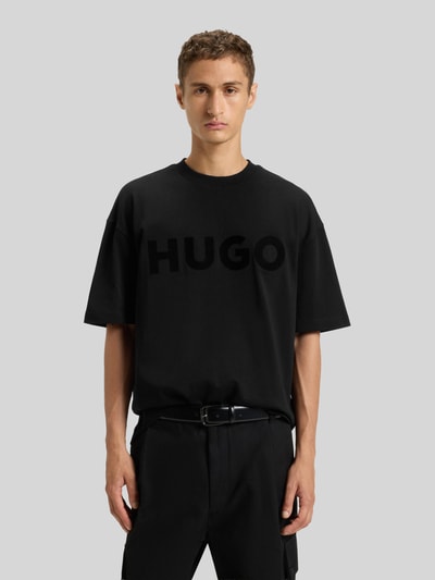 Männermodel trägt Oversized T-Shirt aus reiner Baumwolle Modell 'DINKEE' von HUGO, unifarbenes Schwarz, Label Print, kurzer Arm, Rundhals, Ganzkörperansicht von vorne.