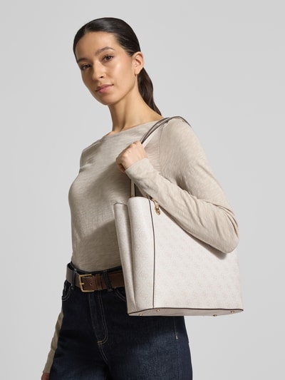 Frauenmodel trägt Handtasche mit Label-Applikation Modell 'NOELLE II' von Guess in Beige mit Logo-Muster und Tragehenkel, Nahaufnahme Oberkörper. Hauptfach mit Reißverschluss, Maße 36 cm Breite, 26 cm Länge, 9 cm Tiefe, Schulterriemen 63 cm.