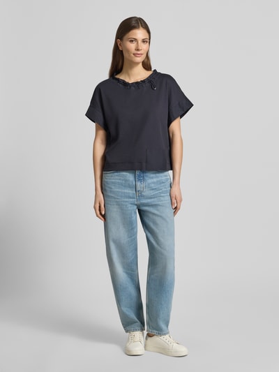 Kobieta nosi T-Shirt mit Eingrifftasche am Saum von Marc Cain, mieszanka bawełny i elastanu, luźny krój, okrągły dekolt z tunelem, wpuszczana kieszeń, pas z tunelem, długość tyłu w rozmiarze 36: 51 cm, ciemny kolor, gładka tekstura, widok całej sylwetki z przodu.