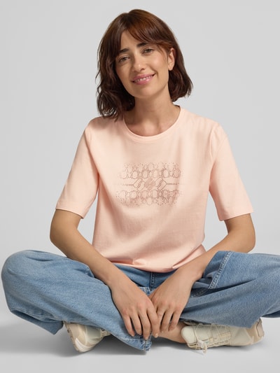 Frauenmodel trägt T-Shirt mit Ziersteinbesatz und Rundhalsausschnitt von Marc Cain, reine Baumwolle, Regular Fit, zarte Rosatöne, Ganzkörperansicht von vorne.