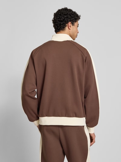 Männermodel trägt Sweatjacke mit Stehkragen von KARL KANI in Braun mit Creme, Material-Mix, Raglanärmel, Label-Stitching, elastische Abschlüsse, Ganzkörperansicht Rückansicht.