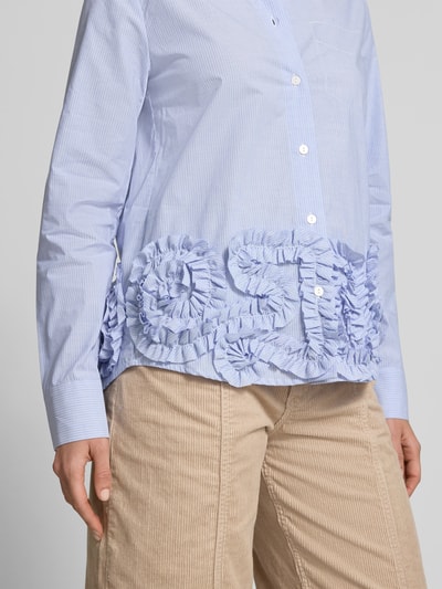 Frauenmodel trägt Regular Fit Hemdbluse mit Rüschenbesatz von BAUM & PFERDGARTEN aus reiner Baumwolle, hellblau-weiß gestreift, durchgängige Knopfleiste, langärmelig, Strukturmix mit auffälligen Rüschen, kombiniert mit beige Cordhose, Nahaufnahme Oberkörper.