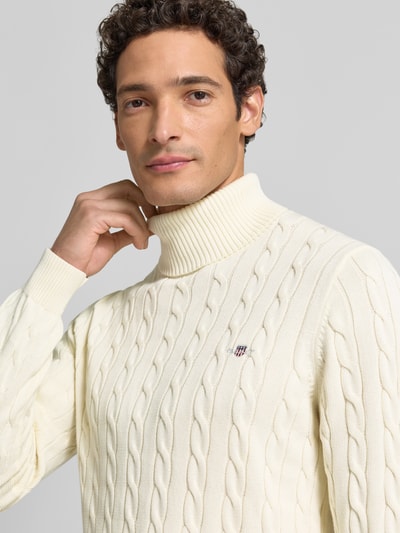 Männermodel trägt Rollkragenpullover mit Logo-Detail von GANT aus reiner Baumwolle, cremefarben, Zopfmuster, Nahaufnahme Oberkörper.