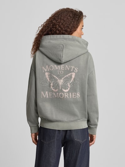 Modelka nosi Oversized Sweatjacke mit Känguru-Tasche Modell 'NIVELLES' von Pegador z mieszanki bawełny, styl oversized, logo, zamek błyskawiczny, kieszeń kangurka, kolor zielonoszary, prosta tekstura, widok całej sylwetki z tyłu.
