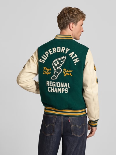 Männermodel trägt College-Jacke aus Woll-Mix mit Eingrifftaschen von Superdry, grün-beige, Stickerei auf dem Rücken, Ganzkörperansicht Rückansicht. Herren College-Jacke von Superdry, Regular Fit, Stehkragen, Knopfleiste, Maße bei Größe M: Rückenlänge 64 cm, Schulterbreite 45 cm, Brustweite 54 cm, Ärmellänge 72 cm.