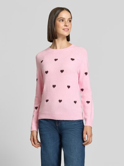 Frauenmodel trägt Regular Fit Pullover mit Rundhalsausschnitt Modell 'DOFFY' von VERO MODA in Rosa mit kleinen dunklen Herzformen, Ganzkörperansicht von vorne. Damen Pullover von VERO MODA