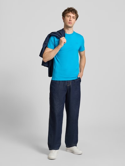 Männermodel trägt T-Shirt mit Rundhalsausschnitt und Label-Stitching von Superdry, reine Baumwolle, Slim Fit, Ganzkörperansicht von vorne.