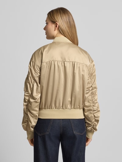 Vrouwenmodel draagt Bomberjacke mit Reißverschlusstasche am Ärmel van Original Bombers in beige, los model met opstaande kraag, gestikt logo, rits, labelpatch, steekzakken met drukknoopsluiting, elastische boorden, ruglengte maat S 49 cm, schouderbreedte 50 cm, borstwijdte 58 cm, mouwlengte 61 cm, gladde structuur, van achteren gefotografeerd, volledige lichaamsweergave rugaanzicht.