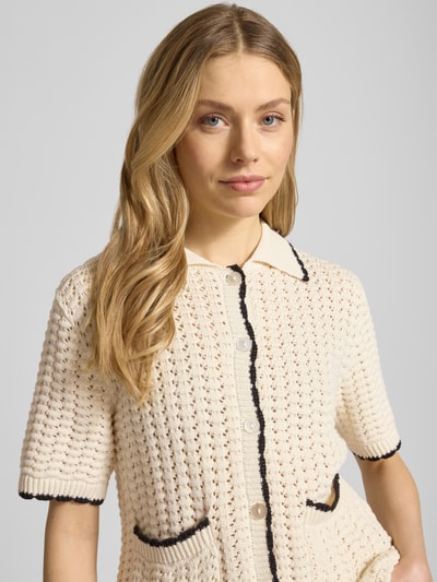 Frauenmodel trägt Regular Fit Cardigan mit Lochmuster Modell 'Muno' von B.Young aus reiner Baumwolle, Strick-Optik, creme mit schwarzen Akzenten, Umlegekragen, aufgesetzte Taschen, Knopfleiste, kurzärmelig, Nahaufnahme Oberkörper.