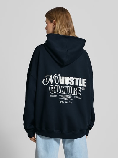 Frauenmodel trägt Oversized Hoodie mit Label-Stitching Modell 'No Hustle' von Karo Kauer aus mieszanka bawełny, czarny, krój oversized fit, kaptur, wyhaftowane logo, kieszeń kangurka, elastyczne ściągacze, tylny nadruk z napisem, Ganzkörperansicht Rückansicht.