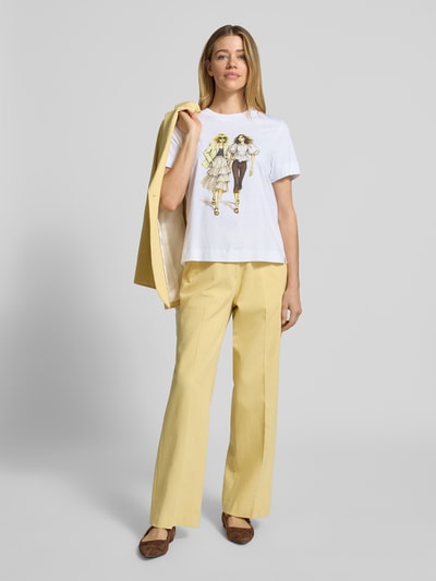 Kobiece model prezentuje damski t-shirt z nadrukiem z motywem i obszyciem kamieniami stras z bawełny ekologicznej marki Rich & Royal, krój straight fit, okrągły dekolt, długość tyłu w rozmiarze S: 58 cm, jasne kolory, gładka tekstura, całopostaciowy widok z przodu.