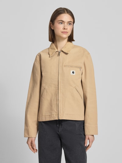 Frauenmodel trägt Jacke mit Label-Detail von Carhartt Work In Progress aus beiger Baumwolle mit Umlegekragen, Eingrifftaschen und Reißverschluss, Relaxed Fit, Ganzkörperansicht von vorne.