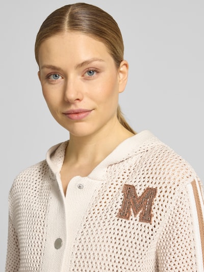 Frauenmodel trägt Regular Fit Strickjacke mit Kapuze von monari in Creme mit Lochmuster, Logo-Detail, Zierbesatz, Nahaufnahme Oberkörper. Reine Baumwolle, AufgesetzteTaschen, Elastische Abschlüsse, Knopfleiste, Rückenlänge 56 cm, Ärmellänge 59 cm, Größe 36.