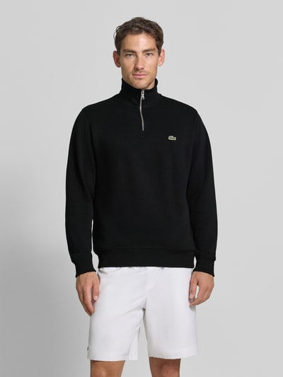 Männermodel trägt Regular Fit Troyer aus reiner Baumwolle von Lacoste in Schwarz, weiße Shorts, glatter Stoff, Ganzkörperansicht von vorne. Herren Troyer von Lacoste