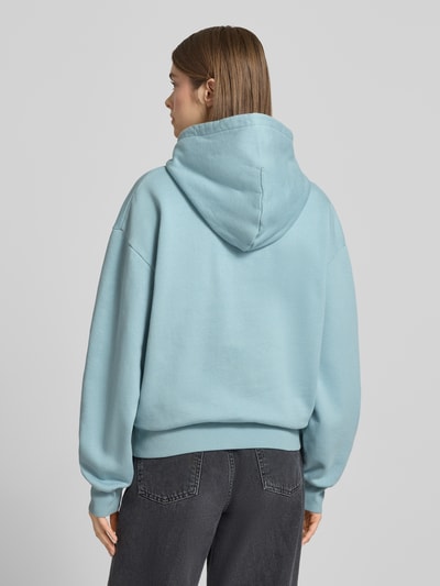 Frauenmodel trägt Hoodie mit Label-Stitching und Känguru-Tasche von Carhartt Work In Progress in Blau, Reine Baumwolle, Relaxed-Fit, Ganzkörperansicht Rückansicht.