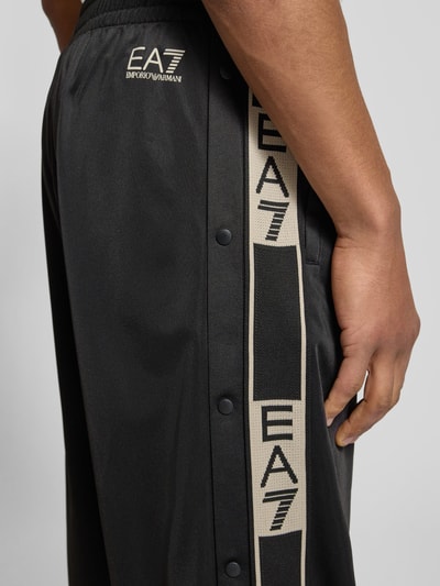 Männermodel trägt Trainingshose mit seitlichen Knopfleisten von EA7 Emporio Armani in Schwarz mit Label-Details, Nahaufnahme Oberschenkel.