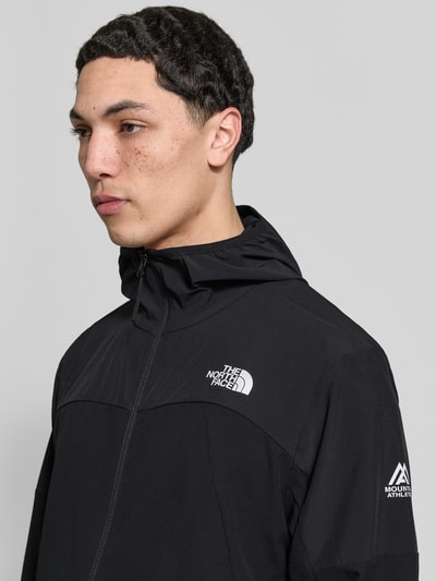 Mężczyzna nosi Jacke mit Logo und Reißverschluss od The North Face, czarna mieszanka materiałów z dodatkiem streczu, logo, kaptur, zamek błyskawiczny, krój regular fit, widok na górną część tułowia z przodu.