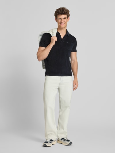 Männermodel trägt Slim Fit Poloshirt aus Frottee von Superdry, kurzärmlig, reine Baumwolle, dunkle Farbe, strukturierte Oberfläche, Ganzkörperansicht von vorne.
