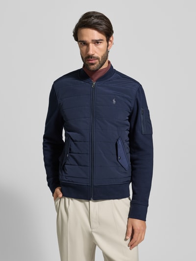 Männermodel trägt Sweatjacke mit gerippten Abschlüssen von Polo Ralph Lauren in Dunkelblau, Regular Fit, Stehkragen, Reißverschluss, Label-Stitching, Pattentaschen mit Druckknopfverschluss, glatte und gesteppte Texturen, Ganzkörperansicht von vorne.