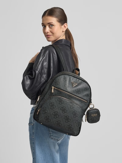 Frauenmodel trägt Rucksack mit Label-Applikation Modell 'POWER PLAY II' von Guess, dunkel mit Logo-Muster und Reißverschluss, Nahaufnahme Oberkörper.
