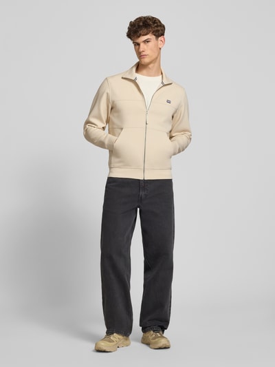 Männermodel trägt Sweatjacke mit Label-Patch Modell 'KOSTER' von Helly Hansen aus Baumwoll-Mix mit Stretch-Anteil in Beige, Gerader-Schnitt, Stehkragen, Reißverschluss und seitliche Eingrifftaschen, Ganzkörperansicht von vorne.