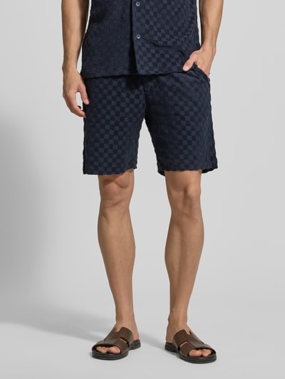 Männermodel trägt Regular Fit Shorts mit Label-Patch von Lindbergh in dunkelblauem Schachbrettmuster, Baumwoll-Mix, Eingrifftaschen, Gesäßtasche, Ganzkörperansicht von vorne.