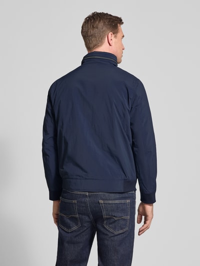 Męski model nosi Regular Fit Jacke mit Stehkragen von Bugatti, granatowa tkanina, zakończenia rękawów elastyczne, zamek błyskawiczny dwustronny, wpuszczane kieszenie na zamek, detal z logo, kaptur chowany, długość tyłu w rozmiarze 50: 65 cm, szerokość ramion w rozmiarze 50: 47 cm, szerokość w rozmiarze 50: 53 cm, długość rękawów w rozmiarze 50: 63 cm, widok całej sylwetki od tyłu