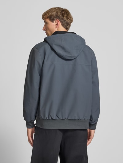 Männermodel trägt Funktionsjacke mit Logo-Stitching und Kapuze von Carhartt Work In Progress, Grautöne, glatte Textur, Ganzkörperansicht Rückansicht. Rückenlänge 70 cm, Schulterbreite 48 cm, Brustweite 55 cm, Ärmellänge 67 cm, gerader Schnitt, Kapuze mit Tunnelzug, seitliche Reißverschlusstaschen, gerippte Abschlüsse.