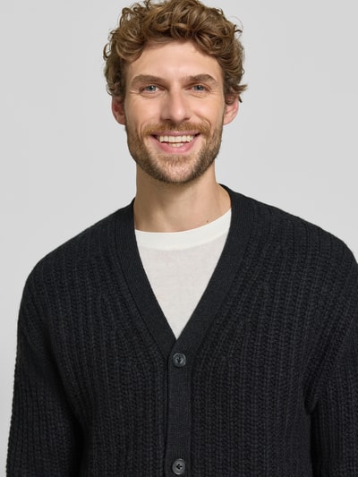 Männermodel trägt Cardigan aus Kaschmir-Mix mit V-Ausschnitt von FTC Cashmere in Schwarz, strukturierte Oberfläche, Nahaufnahme Oberkörper. Gerader Schnitt, Knopfleiste, Stretch-Anteil.