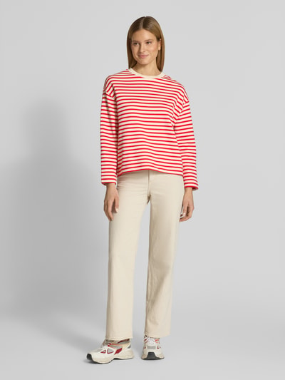 Modelka nosi Oversized Sweatshirt aus reiner Baumwolle Modell 'FRANKAA MAARLEN STRIPE' von Armedangels, dwukolorowa biel i czerwień, długie rękawy, okrągły dekolt, delikatny prążkowany materiał, aplikacja z napisem, luźny krój, widok całej sylwetki z przodu.