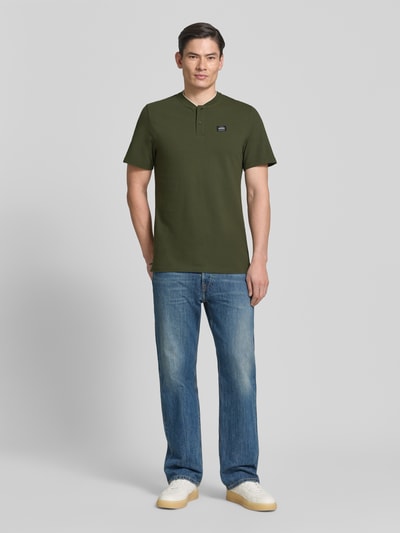 Männermodel trägt Regular Fit Poloshirt aus Baumwoll-Mix von s.Oliver RED LABEL in Dunkelgrün, unifarben, Henley-Ausschnitt, Ganzkörperansicht von vorne.