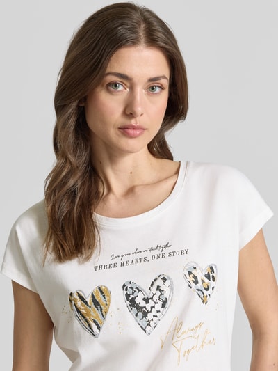 Modelka nosi Regular Fit T-Shirt mit Viskose-Anteil od Christian Berg Woman, biały, nadruk z motywem, bawełniany miks, krótki rękaw, okrągły dekolt, przód z napisem i sercami, krój regular fit, widok na górną część ciała z przodu.