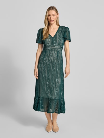 Frauenmodel trägt Kleid mit Häkelspitze von Apricot in dunklem Grün, tailliert, V-Ausschnitt, Volants, leichte Textur, Ganzkörperansicht von vorne.