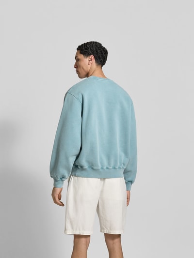 Männermodel trägt Oversized Sweatshirt mit Statement-Print von MAISON KITSUNÉ, hellblau, elastische Abschlüsse, Used-Look, Ganzkörperansicht Rückansicht.