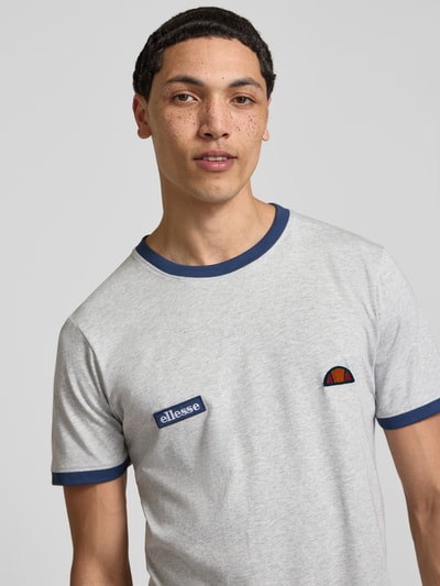 Męski model nosi T-Shirt mit Logo- und Label-Patch Modell 'RINGETTO' von Ellesse, bawełna, krój regular fit, jasnoszary z niebieskimi akcentami, okrągły dekolt, długość tyłu w rozmiarze M: 72 cm, zbliżenie na górną część ciała z przodu.