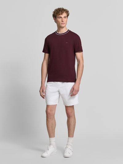 Mannelijk model draagt Regular Fit T-Shirt aus Baumwoll-Mix van Lacoste in bordeaux, witte shorts, sneakers, studio, geheel vooraanzicht.