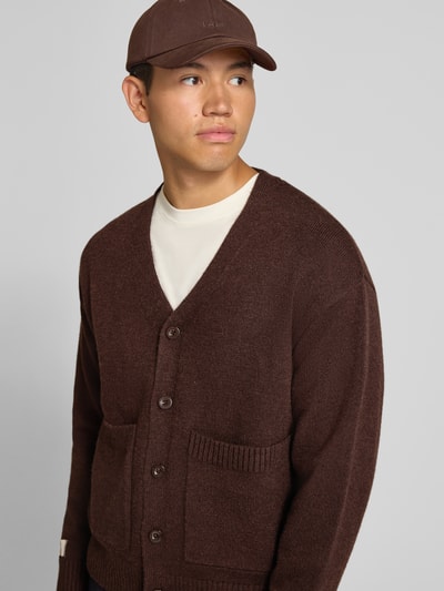 Männermodel trägt Cardigan mit rückseitigem Motiv und V-Ausschnitt von ANOTHER COTTON LAB in Braun, gerade Form, aufgesetzte Taschen, Nahaufnahme Oberkörper.