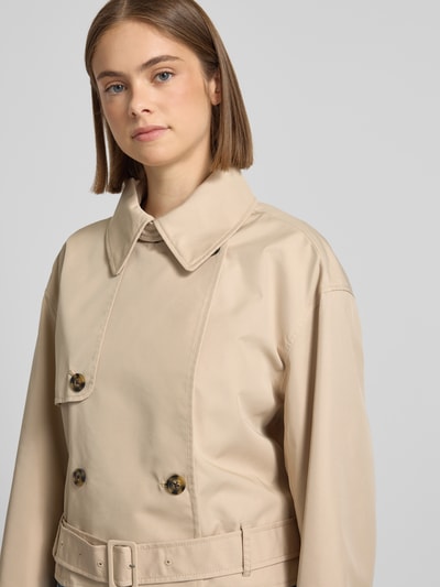 Modelka kobieca nosi Relaxed Fit Trenchcoat aus Baumwoll-Mix od Tommy Jeans, efekt: jednolity kolor, splot twill, mieszanka bawełny, długi rękaw, wykładany kołnierz, listwa guzikowa 2-rzędowa, pasek w talii, naszywka z logo, kieszeń wewnętrzna, skrócony krój, ujęcie: z przodu, zbliżenie na górną część ciała.