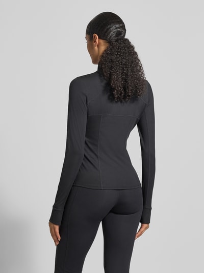 Frauenmodel trägt Slim Fit Jacke mit Stehkragen Modell 'Storm' von Fabletics aus Elastischer-Material-Mix, schwarze Farbe, sichtbare Nähte, Rückenlänge 61 cm, Schulterbreite 34 cm, Brustweite 43 cm, Ärmellänge 66 cm, mit Daumenschlaufen und Kinnschutz, Ganzkörperansicht Rückansicht.