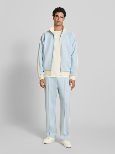 Männermodel trägt Regular Fit Sweatjacke mit Logo-Stitching von KARL KANI in Hellblau mit Stehkragen, Reißverschluss, Eingrifftaschen, Elastischer-Material-Mix, Ganzkörperansicht von vorne.