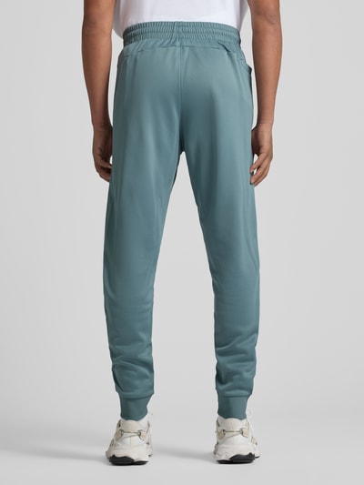 Męski model nosi Sweatpants mit elastischem Bund od Under Armour w luźnym kroju, kolor niebieskozielony, widok całej sylwetki z tyłu. W spodniach elastyczny pas, kieszenie boczne, długość nogawki w rozm. M: 78 cm.