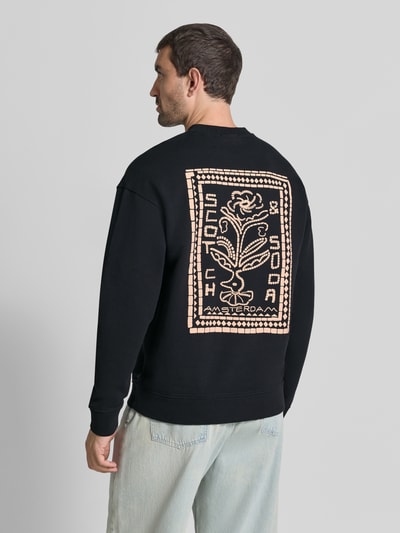Mannenmodel draagt Relaxed Fit Pullover mit Logo-Print van Scotch & Soda in zwart met logoprint op de rug, puur katoen, ruglengte maat M 70 cm, mouwlengte maat M 63 cm, elastische boordjes, pullover met ronde hals, ganzlichaamsaanzicht achterkant.